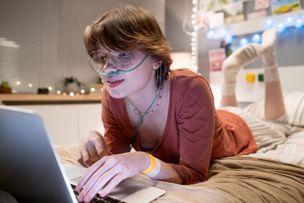 Cute brunette teenager lying on bed in front of laptop screen while networking or communicating in video chat